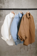 Three shirts in white, blue, and brown hanging on a rack against a concrete wall.